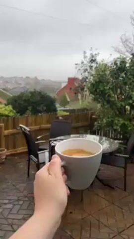 The video shows a hand holding a grey mug full of coffee outdoors on a rainy day. The mug is then placed on a glass-topped table surrounded by four chairs on a brick patio. A low wooden fence separates the patio from a yard with various shrubs. In the distance, a neighborhood of houses is visible against an overcast sky. Raindrops are visible on the table and the patio.