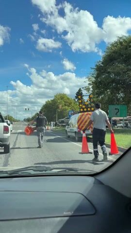 The video shows a scene from inside a car, looking out through the windshield. In the foreground, the car's dashboard is visible. Through the windshield, two men are walking across the road, carrying orange traffic cones. The man on the left is wearing a camouflage-patterned uniform with a dark vest, while the man on the right is wearing a light grey shirt and dark pants with reflective stripes. 

A white van with yellow and red stripes is parked on the side of the road, featuring lights on top. The van has the word "SEGURIDAD VIAL" written on its side. There's a traffic sign nearby with the number "140" and an image of a telephone. In the background, there are other cars, trees, and a traffic light. The sky is blue with white clouds.