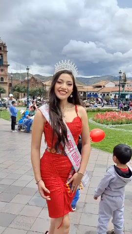 A young woman wearing a red dress and a crown stands in a public square. She is also wearing a sash that says "REINA CUSCO CIUDAD". She is smiling and holding a red balloon. In the background, there are people, buildings, mountains, and a cloudy sky.