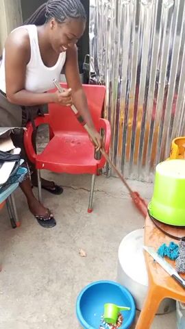 A smiling, dark-skinned woman with braided hair is sweeping a concrete floor in a small outdoor space. She is wearing a white tank top and brown shorts, and flip-flops. She uses a red broom with a silver handle, sweeping around a red plastic chair. To the left is a small, colorful table with items on it, including a black jacket. To the right is a small table with a green bucket and other items. The background consists of corrugated metal walls.