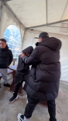 In a tent-like structure, three individuals, each wearing puffy jackets, are present. Two of them engage in what appears to be roughhousing. One person initially tries to grab the other, but the latter turns and pushes the first individual away.  Meanwhile, the third person watches the scene. A wooden table and benches are visible, placed in the center of the tent, which has a white ceiling and a window with a view of the outdoors. Subsequently, a young man looks into the camera.  The camera then focuses on someone sitting, facing the benches. The overall ambiance is one of casual playfulness.