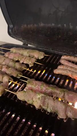 The video shows an open grill with chicken skewers and sausages cooking. The chicken skewers are arranged on the left side of the grill, and the sausages are on the right. Flames are visible beneath the grill grates. The grill is set against a dark background.