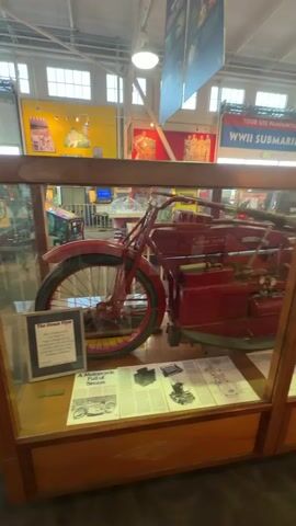 The video pans across a display case containing a vintage red steam-powered motorcycle. The motorcycle is prominently featured, with its front wheel, engine, and rear wheel visible. The display case is made of wood and glass, and inside, there are various informational materials displayed, including what appear to be diagrams and newspaper clippings about the motorcycle. In the background, the interior of a museum or exhibition hall is visible, with other displays and signage, including one that reads "WWII SUBMARINE." The lighting suggests an indoor environment.