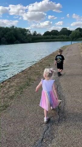 A young boy and girl are walking along a paved path next to a body of water. The girl is in front, wearing a colorful dress. The boy is behind her, wearing a black shirt and shorts. The path is lined with grass and trees. The sky is blue with white clouds. The water is rippling.