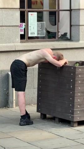 A man is seen outdoors in what appears to be a city, leaning on a dark wood planter. He is shirtless and wearing black shorts and black shoes. His head and arms rest on the top of the planter. Behind him is a window with advertisements on it. The ground is paved.