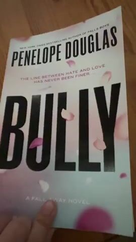 The video shows a person flipping through a book with a wooden floor in the background. The book appears to be a paperback with a pink and black cover. The title is partially visible as PENELOPE DOUGLAS. The pages of the book are white with black text. The person's hand is visible as they flip the pages. The lighting is natural and appears to be coming from the side. The scene is close-up and focused on the book.