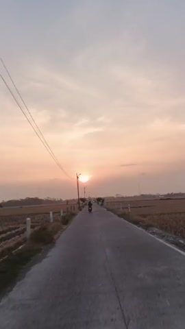A motorcycle is seen riding down a paved road in a rural area. The sun is setting, casting a warm glow over the sky and landscape. Fields of dry crops flank the road, and power lines stretch overhead. The overall scene is peaceful and idyllic.