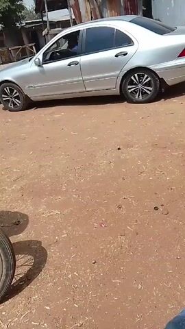 The video shows a person sitting on a motorcycle. The motorcycle wheel is visible in the foreground. A silver sedan is parked to the side on a dirt road. In the background are tin buildings. The person is wearing blue jeans and black shoes.