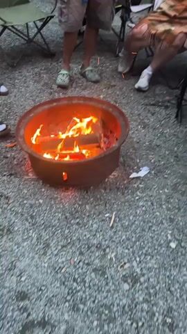 The video shows a close-up, overhead shot of a burning fire pit surrounded by gravel. Several people are visible in the background, seated or standing around the fire pit. The fire pit is round and made of a reddish-brown metal. It contains several logs that are actively burning, producing bright orange and yellow flames. The people in the background are wearing casual clothing, including shorts and sandals. Some are sitting in folding chairs. The overall environment appears to be an outdoor setting, possibly a campsite, given the presence of the fire pit and chairs. The lighting suggests it is either dusk or overcast.