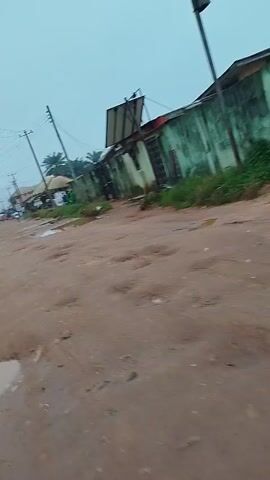 The video shows a muddy dirt road with several large potholes. On the right side of the road is a dilapidated green building with a corrugated metal roof. Some people are visible in the distance near the building. The sky is overcast, suggesting it may have recently rained or is about to rain. The overall scene appears to be in a rural or impoverished area.