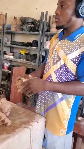 A man is standing in a pottery studio, wearing headphones and a colorful shirt with the name “ASHWAN” on it. He is kneading a lump of brown clay in his hands. A second lump of clay sits on a slightly messy worktable. The clay has been partially shaped, appearing almost skull-like. Behind the man, shelves are filled with various pottery pieces, some covered in black plastic. The studio appears to be well-used and functional. The room is well-lit with natural light.