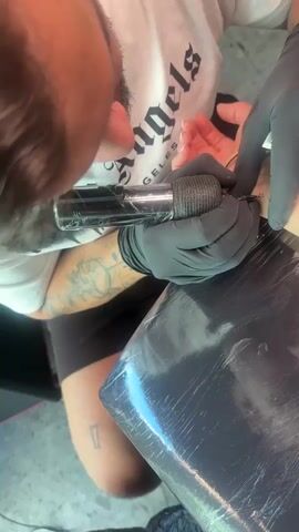 This close-up video shows a tattoo artist in black gloves holding a tattoo gun wrapped in black grip tape and applying a tattoo to the wrist of a person. The tattoo artist's face is visible in the upper left corner of the frame, and they are wearing a white t-shirt with the word "Angels" on it. The person receiving the tattoo has their arm resting on a surface covered with black plastic wrap. Other visible details include tattoos on the artist's arm and a ring on the recipient's finger. The video is oriented vertically.