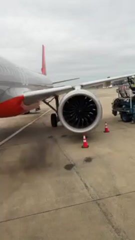 From a low vantage point on an aircraft boarding bridge, we observe a jet engine, likely on an Airbus A320 series aircraft given its general configuration. The engine is running, producing a visible exhaust plume near the ground. The aircraft has a grey fuselage with red accents. Two orange traffic cones are placed in front of the engine. Behind the engine, ground crew and equipment are visible. The weather appears overcast. The camera briefly pans upwards to show the boarding bridge structure.