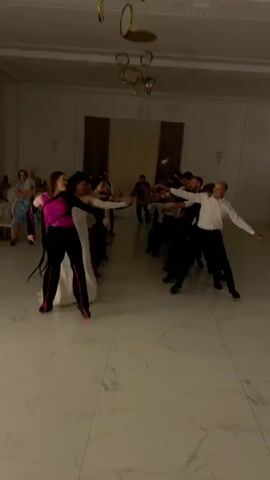 A group of people are dancing in a well-lit ballroom. The floor is white marble with a subtle pattern. In the foreground, a woman in a pink top and black pants dances with a man in a white shirt and black pants. They are facing each other with their arms extended. Behind them, more people are dancing in two lines, facing each other. Some are dressed formally, while others are in casual attire. The room has high ceilings with a decorative chandelier. In the background, tables are set up, and some guests are seated, observing the dancers. The lighting in the room changes throughout the video, with moments of bright white light, red light, and spotlights.