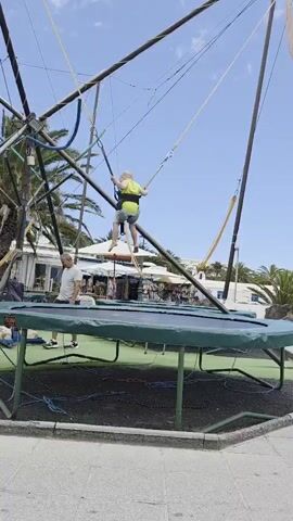 A child in a yellow vest and grey shorts is bouncing on a bungee trampoline. The child is suspended in the air by elastic cords attached to a harness. The trampoline is green and has a metal frame. A man in a white t-shirt and dark shorts is standing on the ground next to the trampoline, watching. In the background, there are palm trees, white buildings, and a clear blue sky with some clouds, indicating it is daytime. The scene appears to be an outdoor amusement area or park in Costa Teguise, Spain, possibly a tourist resort given the presence of palm trees and beach-like setting. The activity is recreational, suggesting a fun and energetic atmosphere. The child's posture implies movement and height achieved through bouncing.