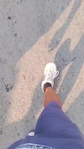 The video begins with a close-up shot of a person's feet walking on an asphalt surface, casting long shadows. The person is wearing white sneakers and dark blue pants. The camera then pans up to show a group of people walking along a paved road. The group appears to be heading in the same direction, with individuals in various clothing, including t-shirts and pants. Some people are wearing white shirts with numbers, suggesting they might be part of a team or event. There are shadows of the people on the road, indicating it's daytime with the sun at an angle. Further down the road, there are buildings and trees visible on the sides. Several motorcycles are also seen on the road, with one appearing to be carrying a rider. The overall scene depicts a group of people walking together outdoors on a sunny day, possibly in a procession or moving through a populated area.
