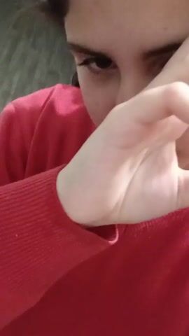 A person is wearing a red long-sleeved shirt and partially obscures their face with their hand. Only one eye is visible. The background is a blurred, gray wall.