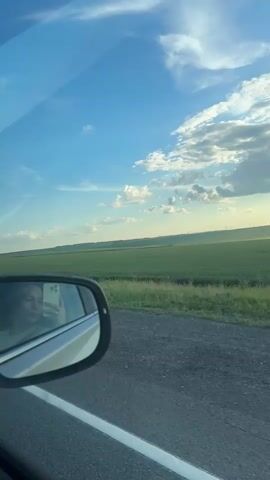 A woman is taking a selfie in a car's side mirror while a green field and blue sky pass by. The video is shot from inside the car, showing the road and the passing scenery. The woman appears to be wearing a light-colored shirt. The sun is visible in the distance, casting a warm glow on the landscape. The overall mood is peaceful and scenic.