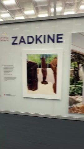 The video shows a person walking past displays of artwork. The titles on the wall read "MUSÉE ZADKINE" and "MUSÉE DE LA VIE ROMANTIQUE". There are framed pictures on the wall and people walking in the distance down a long hallway. The building has a very high ceiling with a cross-hatched pattern.