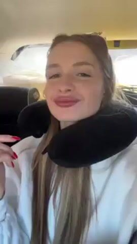A young woman wearing a white hooded sweatshirt and a black travel neck pillow is filming herself in the back seat of a car. She has long hair parted in the middle, and her nails are painted red. She smiles and sings along to music playing in the car. The background is slightly out of focus, showing a car interior and glimpses of the outside.