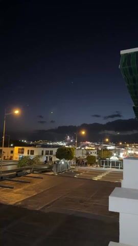 The video shows a nighttime cityscape with fireworks. The view is from a rooftop, overlooking a town or city with white buildings and lit street lamps. The sky is dark, with some clouds visible. Fireworks explode in the distance, with red and gold colors.