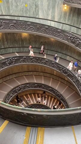 The video presents a high-angle view looking down into a grand, multi-level spiral staircase within the Vatican Museums, Città del Vaticano, Vatican City. Numerous individuals are seen walking down the expansive, winding stairs. The staircase features elaborate, dark-colored wrought iron railings with decorative elements, contrasting with the lighter steps and the green and white marble-patterned walls that curve along the inner and outer perimeters. Yellow lines are visible on the ground level at the bottom of the staircase, along with a yellow triangular warning sign on the upper left wall. The overall impression is one of architectural grandeur and steady, leisurely movement of people through a significant historical or cultural site.