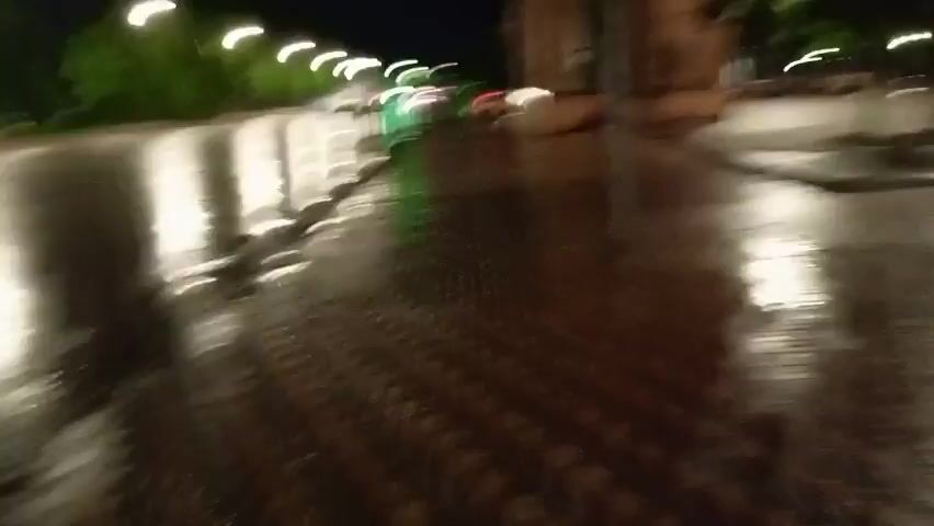 A first-person perspective video shows a wet, paved walkway at night. Streetlights cast long, bright reflections on the wet surface. There are trees and some buildings visible. A few people are walking in the distance. There appears to be a fountain in the middle ground. The camera is moving forward.
