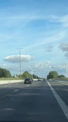 This is a vertical video clip of a car driving on a highway in Perwez, Belgium. The scene features a two-lane highway with a white dotted line separating the lanes and a solid white line along the shoulder. Several cars are visible on the road, including a black SUV in the foreground, a white car and a dark sedan further ahead.

The surrounding landscape is characterized by lush green trees and fields on either side of the highway. Dominating the background are numerous large white wind turbines, some actively spinning their blades, suggesting a breezy day. The sky is a bright blue with scattered fluffy white clouds. Tall lampposts are also present along the roadside. The overall impression is one of a journey through a scenic, modern landscape powered by renewable energy. The time of day appears to be daytime, given the bright sky and clear visibility. There are no discernible emotions or specific events beyond the act of driving. No visible text is present in the clip.
