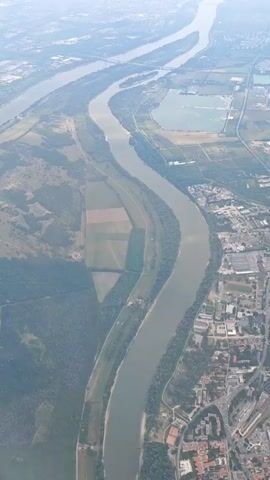 This is an aerial view video of a wide, meandering river with a city situated on its right bank. The river is the dominant feature, with green, forested islands and agricultural fields visible on its banks and within its curves. The city, characterized by a dense collection of buildings and infrastructure including roads, train tracks, and industrial areas, extends along the river. The overall environment appears to be a mix of natural landscape and urban development under a hazy sky. The camera angle is high and angled downwards, giving a sweeping perspective of the landscape. There are no people or specific actions clearly discernible due to the altitude and distance.