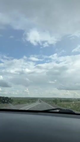The video displays a first-person view from inside a moving vehicle, looking forward through the windshield. The vehicle's dashboard is visible at the bottom of the frame. A straight asphalt road stretches into the distance, flanked by green fields or grass under a partly cloudy sky with patches of blue. White road markings are visible on the asphalt. A vehicle can be seen driving away in the distance. As the camera approaches, two traffic signs, one red 'no entry' and one blue circular sign, become visible on the left side of the road. The windshield has small droplets or particles on its surface.