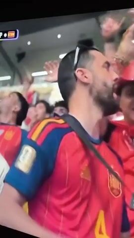 This is a video showing a soccer game from the perspective of a spectator. The game is between Portugal and Spain, as indicated by the score displayed on screen. In the foreground, a man wearing a red Spanish soccer jersey with a gold FIFA World Cup patch on his left arm is visible. He is looking towards the field. In the background, the soccer field is shown with players in red and white uniforms. The stadium is filled with spectators. The game appears to be in progress. The man in the foreground is wearing sunglasses on top of his head.