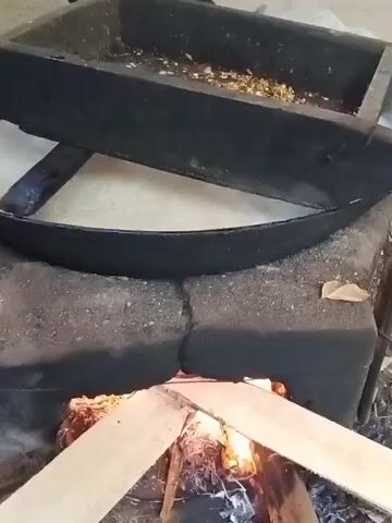 A slow-motion video shows a black pan with some lighter-colored material in the bottom, resting on a curved metal frame, which in turn is on top of a stone structure with an open fire burning inside. The fire is fueled by logs and is producing smoke. The pan is positioned to be heated by the fire. The surrounding environment is indistinct but appears to be outdoors or in an open-air setting.