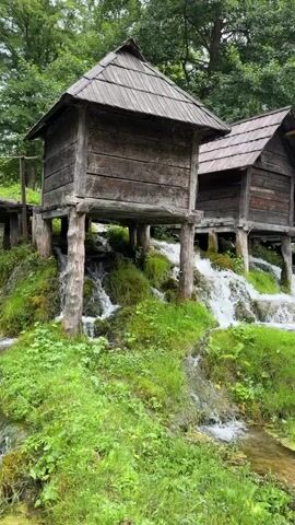 The video shows a series of old, wooden huts built on stilts over a cascading stream. The huts have weathered wooden walls and shingled roofs. The stream flows over moss-covered rocks, creating a series of small waterfalls. Lush green grass and trees surround the scene, and the overall environment appears to be natural and serene. The camera pans slowly from left to right, showcasing multiple huts and the surrounding landscape. There are no people visible in the immediate foreground, but a blurred figure can be seen on a path in the distance.