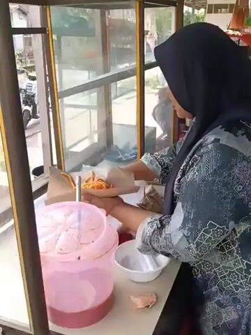 A woman wearing a dark hijab and a patterned shirt is standing behind a glass-enclosed food stall. She is holding a skewer of food in her right hand and appears to be placing it into a cone-shaped paper wrapper with her left hand. In front of her on the counter is a pink plastic container with a patterned lid, a small white bowl, and a piece of food. The background shows a street scene with a white motorcycle parked on the left and blurry figures of people in the distance. The stall has a wooden frame and appears to be outdoors.