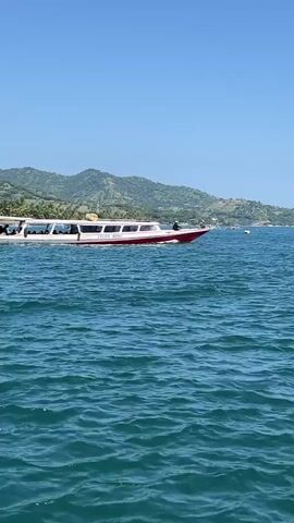 The scene features a bright, clear day with a tranquil sea under a cloudless blue sky. In the foreground, a boat with a red and yellow hull passes by. Further off, a larger white boat with a red stripe along its hull is moving across the water, transporting several passengers. In the background, a coastline with green hills and trees is visible. The overall impression is one of a peaceful, sunny day at sea.