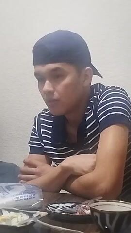 A young man is sitting at a table with his arms crossed. He is wearing a navy blue baseball cap and a navy and white striped polo shirt. He looks at the camera. There is a plate of food, a cup, and a package of napkins on the table in front of him. The background is a plain white wall.