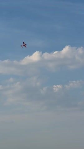 A small, white and red airplane is captured in mid-flight against a backdrop of a bright blue sky dotted with fluffy white clouds. The airplane, which appears to be a model or remote-controlled aircraft, is angled as if banking or turning, with its wheels visible. The camera is positioned below the aircraft, looking upwards, emphasizing its aerial movement. The sunlight suggests a daytime setting, and the clear visibility indicates fair weather conditions. There are no people visible in the frame.