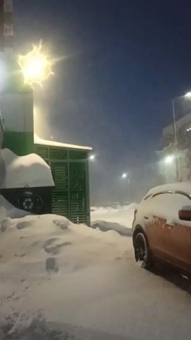The video shows a snowy scene during what appears to be the early evening or late afternoon. Snow is falling heavily, partially obscuring the view.  An orange car, partially covered in snow, is parked on the right. Next to the car is a green structure which is also laden with snow.  A street light is visible above and behind the structure.  In the distance, beyond the structure, what looks like a parking lot with additional snow-covered vehicles is visible.  A streetlight is also visible in the distance. The sky is a dark blue.