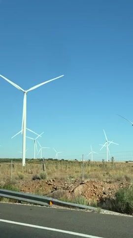 The video shows a view from a vehicle traveling along a road. In the foreground, there is a gray metal guardrail and the edge of an asphalt road with white markings. Beyond the guardrail, the landscape consists of dry, scrubby vegetation and some green bushes. Dominating the background are numerous white wind turbines, some with their blades spinning, set against a clear blue sky. The wind turbines are spread across a gentle hillside. A blue traffic sign with two white upward-pointing arrows is visible on a pole to the right of the frame for a brief moment. The scene appears to be a rural area with a focus on renewable energy production.