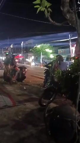 A red tanker truck with "PERTAMINA" written on its side is parked on a street at night. Several people are gathered around the truck, some sitting on the ground. A motorcycle is parked in the foreground. Lights are visible in the background, suggesting a populated area. The scene appears to be outdoors.