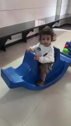 A young child with dark curly hair, wearing a white long-sleeved shirt, brown shorts, and brown shoes, stands on a blue plastic rocking boat toy in a room with light tile flooring. The child is holding onto the side of the boat for support, smiling and looking around. Behind the child is a long black bench against a wall with light-colored tile and a dark trim. Other colorful toys are partially visible near the rocking boat.