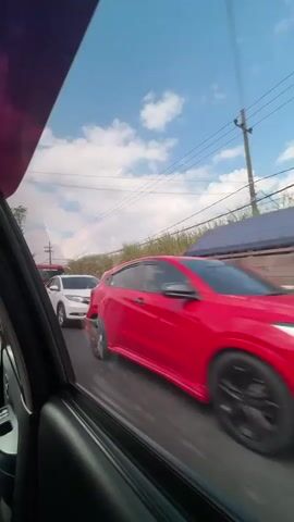 A side view from inside a vehicle shows a line of traffic on a road. A white SUV is in the left lane, followed by a red car with black rims in the right lane. A red tow truck with motorcycles loaded on its flatbed is in the left lane, passing the white SUV. A black SUV is in the left lane, followed by a motorcycle. Tall, dry grass is visible on the right side of the road, and power lines are strung across the sky. The sky is blue with scattered white clouds.