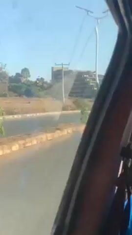 The video shows a view from inside a vehicle, looking out at a multi-story building on the side of a road. The building appears unfinished or derelict, with visible concrete and irregular levels. The sky is clear and blue. There are light poles and greenery along the roadside. A second vehicle can be seen on the road. The video appears to be taken while in motion.