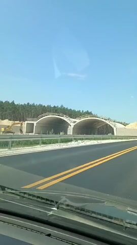 This is a dashcam video recorded from the perspective of a vehicle driving on a road in Mała Nieszawka, Poland. The video shows a sunny day with a clear blue sky. The primary subject of the video is a bridge or overpass under construction. The structure features two large arched openings made of concrete. Some sections of the bridge are still covered with scaffolding and protective sheeting, indicating ongoing construction work.

In the foreground, the road is paved and marked with double yellow lines, suggesting it is a main thoroughfare. The vehicle passes by a large white semi-trailer truck with red and gold lettering. The visible text on the truck includes "KNUTH" and "040 / 768 68 49," likely a company name and phone number. The truck is in motion, traveling in the same direction as the recording vehicle.

As the vehicle approaches and passes through the arched openings of the bridge, other vehicles, including cars and possibly another truck, can be seen on the road ahead and within the structure. The surrounding landscape appears to be rural, with a line of coniferous trees visible beyond the construction site. The overall mood of the video is neutral, simply documenting a drive on a highway with ongoing infrastructure development.