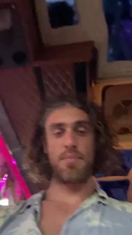 A man with curly brown hair and a beard is looking up. He is wearing a light blue short-sleeved shirt with a floral pattern. The background appears to be the interior of a vehicle, possibly a bus or RV, with padded upholstery on the ceiling and some colorful lights.