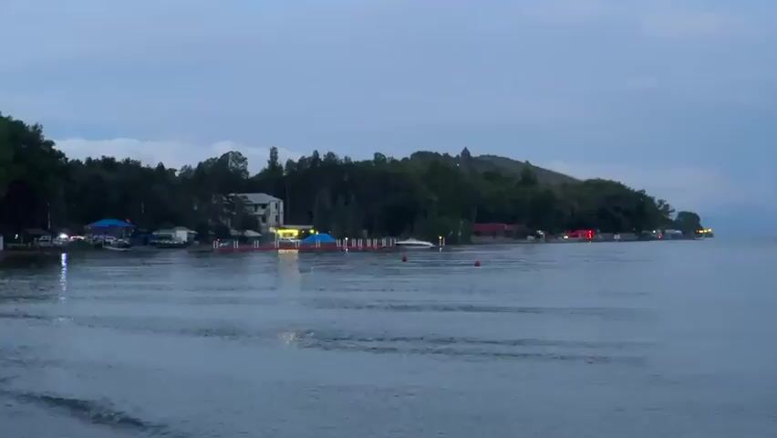 The video shows a wide shot of a body of water, likely a lake or ocean, with a shoreline lined with trees and buildings. The time appears to be dusk or dawn, with a cloudy sky casting a dim light over the scene. Several boats are visible docked along a pier on the left side of the frame, with some lights illuminated on the shore. In the distance, a few red buoys are floating on the water. The camera pans slightly to the right, revealing more of the shoreline and the vast expanse of the water under the muted sky. There are no people clearly visible in the frame.