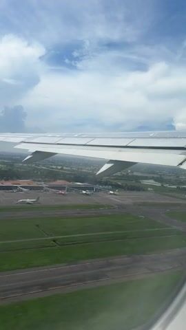 A daytime aerial shot from an airplane window captures the aircraft's wing and the landscape below. The prominent white wing occupies the upper half of the frame, with its flaps extended downwards, indicating either takeoff or landing. Beneath the wing, an airport is visible, featuring asphalt runways, taxiways, and several parked aircraft of various colors.

The surrounding area consists of vibrant green fields, intermixed with numerous rectangular bodies of water, likely rice paddies or aquaculture ponds, suggesting a rural or semi-rural setting. Scattered buildings and structures are also visible within the landscape. The sky above is partly cloudy, with significant patches of blue sky and large white cumulus clouds.

The perspective suggests the plane is in the process of ascending, moving away from the airport. No specific text is visible in the frame. The sound in the video corresponds to an active airplane engine, consistent with takeoff or initial climb.
