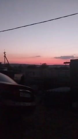 A car is parked in the foreground, partially obscured by darkness. In the background, a sunset paints the sky with shades of pink and purple. Silhouetted against the sky are power lines, a utility pole, and the faint outlines of buildings and trees. The scene appears to be a rural or suburban setting at dusk.