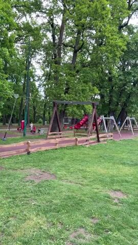 The video shows a playground in a park on a cloudy day. The playground features swings, slides, and geometric climbing structures.  People, including children and adults, are seen walking around the playground and engaging with the equipment. The ground is covered with grass and paved pathways. There are also some bicycles parked near one of the climbing structures. Tall green trees surround the playground, and power lines are visible overhead.