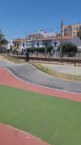 A person is seen skateboarding on an outdoor pump track under a clear blue sky in Premià de Mar, Spain. The skateboarder, wearing a blue t-shirt, dark pants, and a dark cap, navigates the gray asphalt track which features various ramps, bowls, and banked turns. Sections of red and green colored ground border the track.

The skateboarder skillfully maneuvers through the track, pumping to gain momentum and riding up the curved walls of the bowls. Palm trees and multi-story buildings, typical of a coastal town, are visible in the background. In the distance, a sandy beach and the sea can be seen, along with a city skyline. A white boat is partially visible on the left side of the frame.

On a low white wall adjacent to the track, the word "BRAVADO" is visible in black lettering, along with a red "STOP" sign. The scene is brightly lit by the sun, indicating it is daytime. The skateboarder appears focused on their ride, demonstrating control and agility on the track.